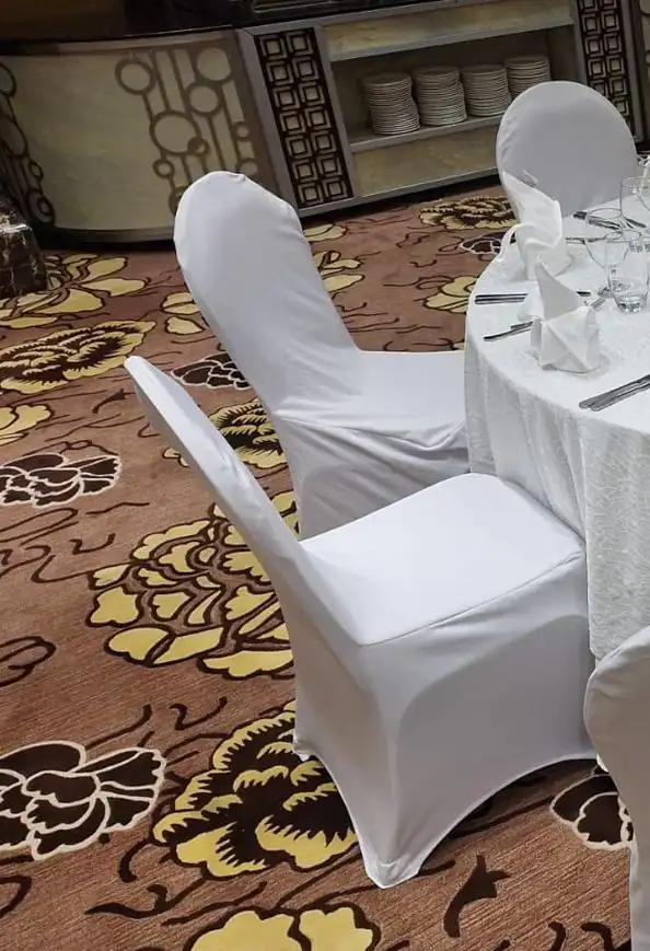 طاولة مستديرة بمفرش أبيض وكراسي مكسوة بأغطية بيضاء فوق سجاد مزخرف