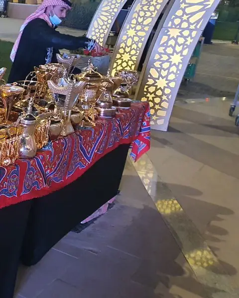 طاولة بوفيه بمفرش أحمر مزخرف عليها دلال قهوة عربية في حديقة